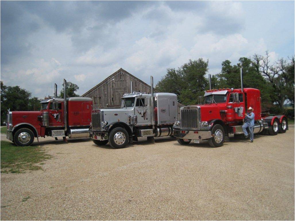 Three Evans Ranch Peterbilts parked in a row on the gravel in front of the old Wilson Valley barn, one of the drivers standing by the red truck on the right