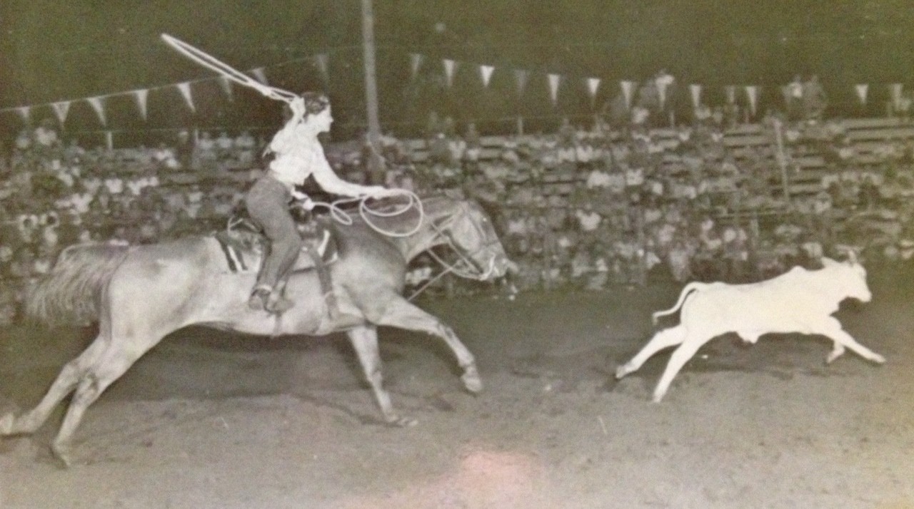 Bernice Evans roping a calf on horseback in a rodeo arena, the grandstand full behind her.