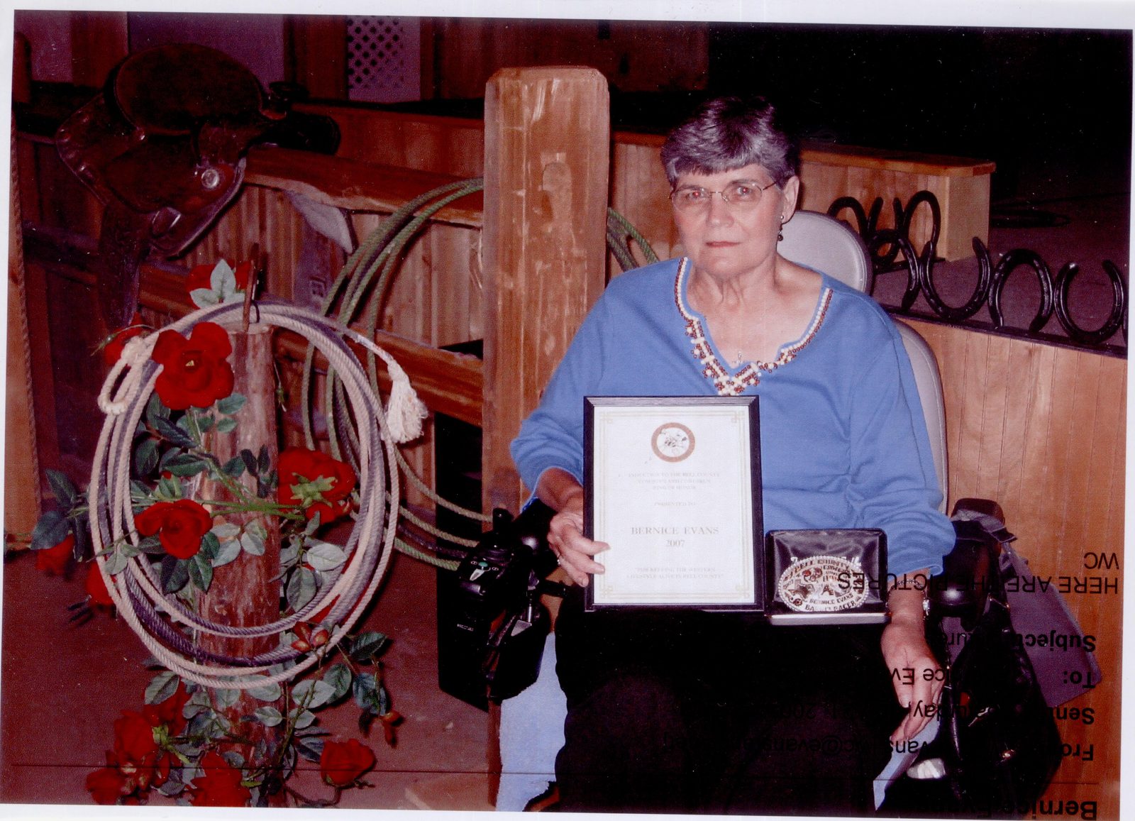 Bernice holding a framed certificate and a buckle beside a display of ropes and horseshoes, her cowgirl award.