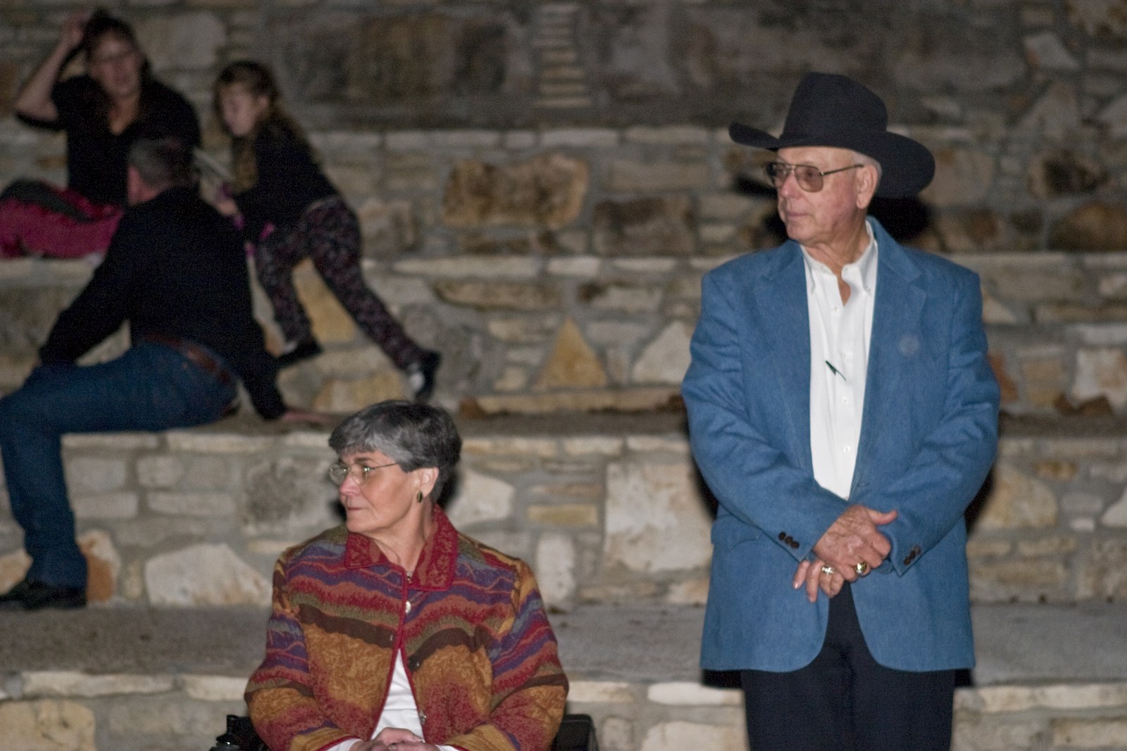 W.C. standing in a blue jacket and black hat, Bernice seated on the stone wall in front of him, at John and Erica’s wedding.