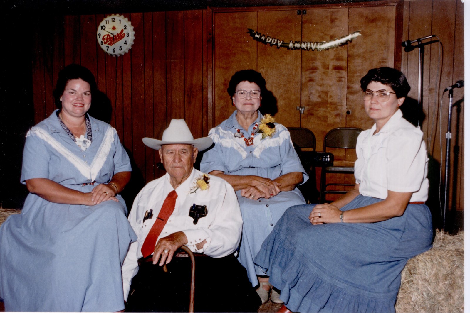Three women in blue and an older man in a white shirt and hat seated in a row at a Jones anniversary.