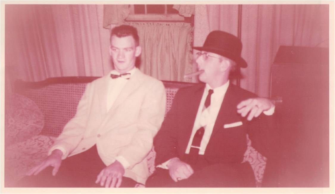 Two young men in suits and bow ties on a couch, an old colour print.
