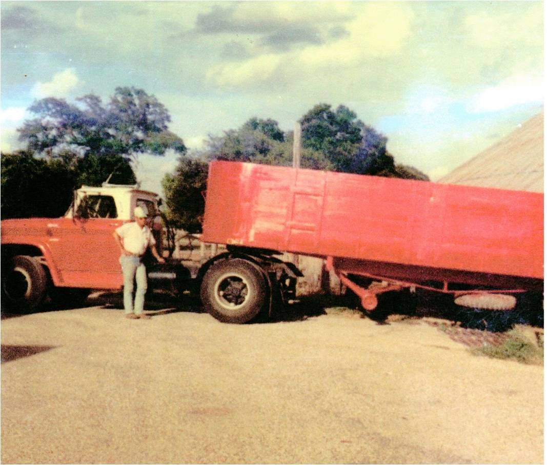 A boy standing by a red grain truck with its bed tipped up.