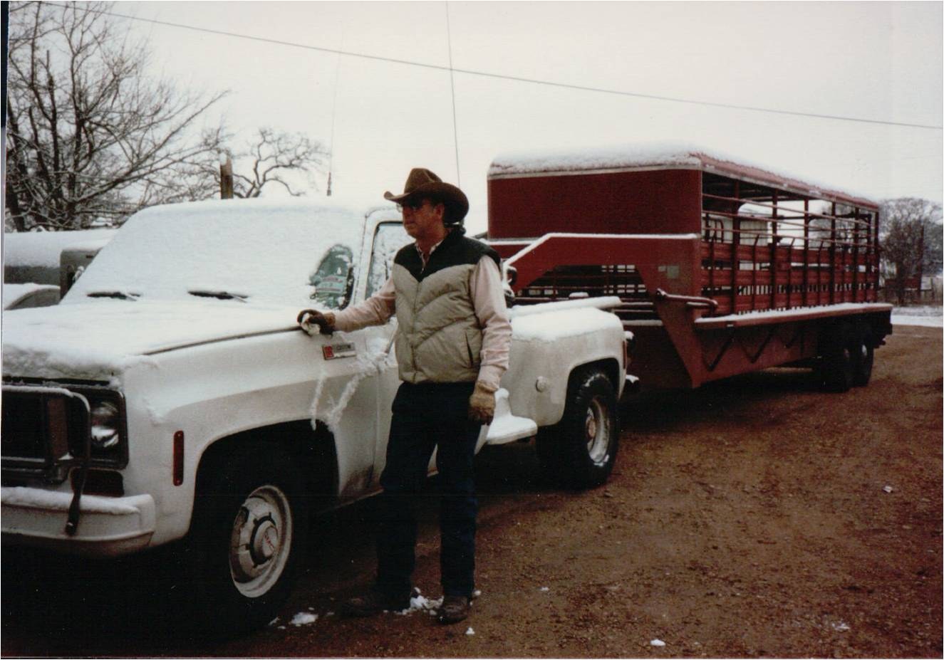 A man in a hat beside a snow-covered white pickup hitched to a cattle trailer.