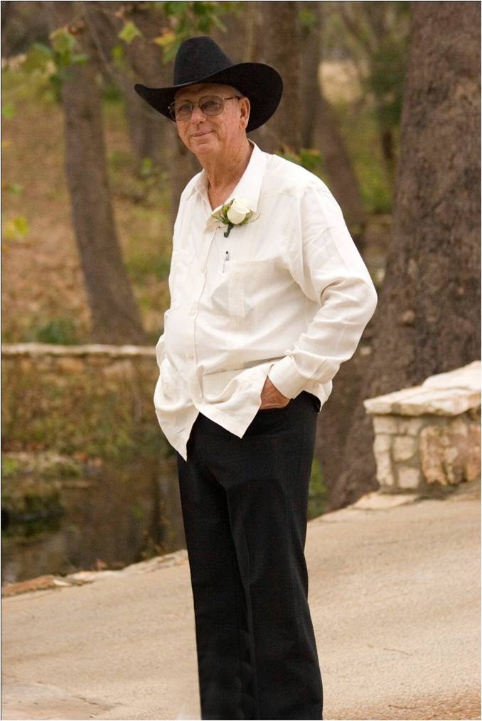 A man in a black hat and white shirt with a boutonniere, standing among the trees.