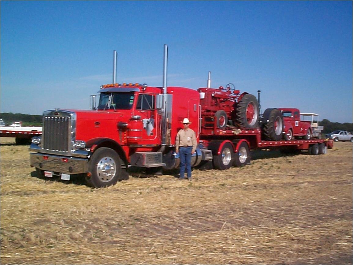 A man in a white hat in front of a red Peterbilt hauling a red tractor on a lowboy.