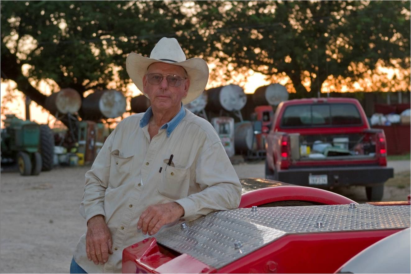 A man in a white hat leaning on a red pickup at sunset, farm equipment behind him.