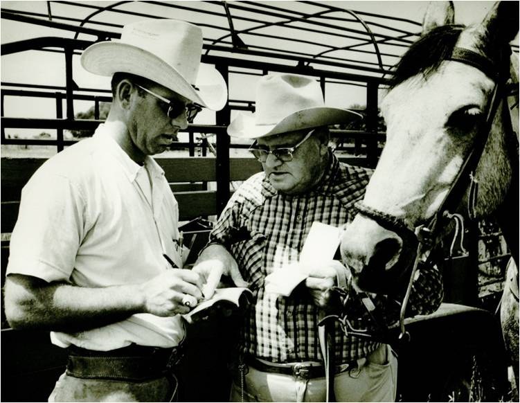 Two men in hats at a horse’s head by the pens, one writing on a clipboard. Black and white.