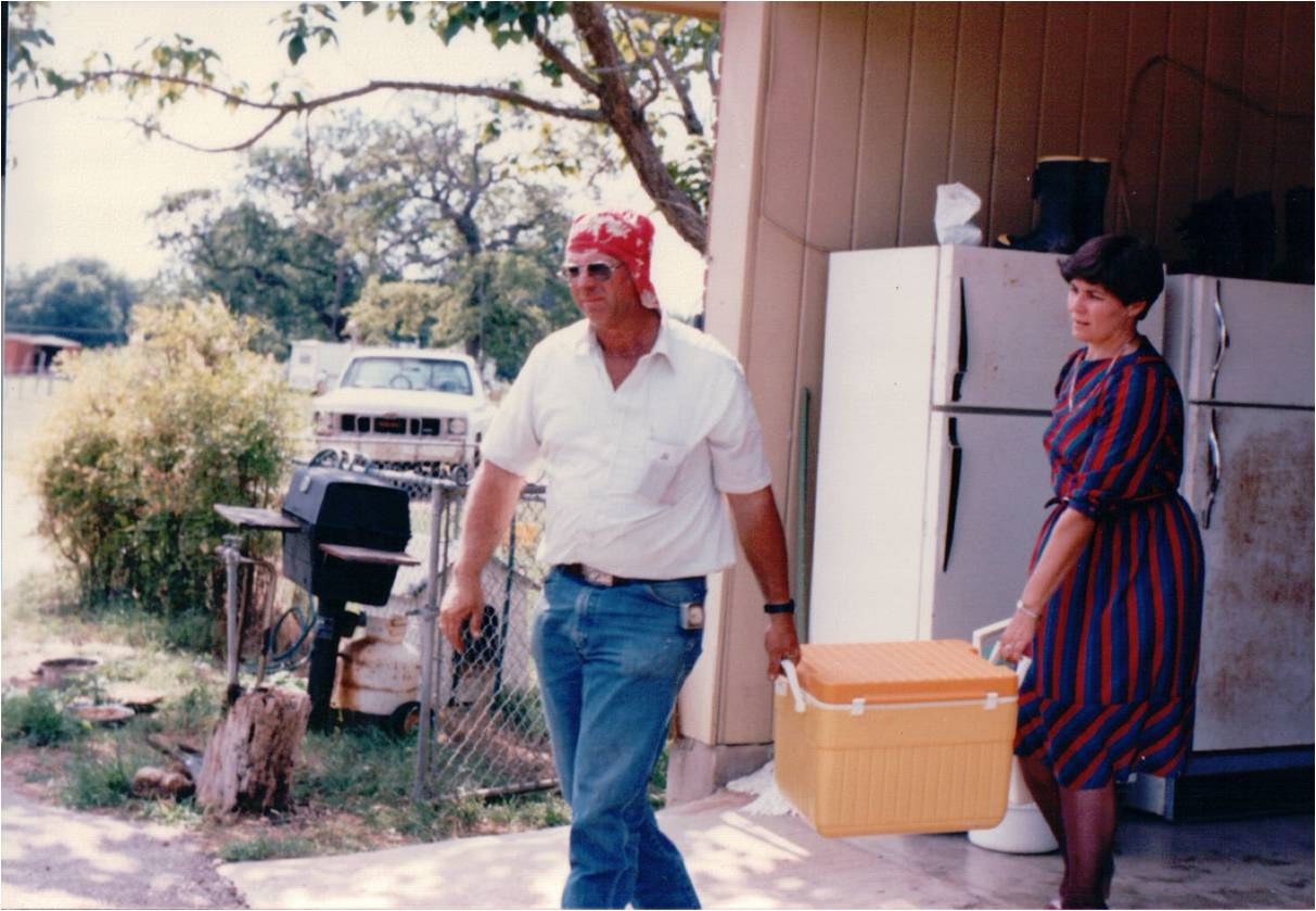 A man in a red cap carrying a cooler and a woman in a striped dress by the shed.