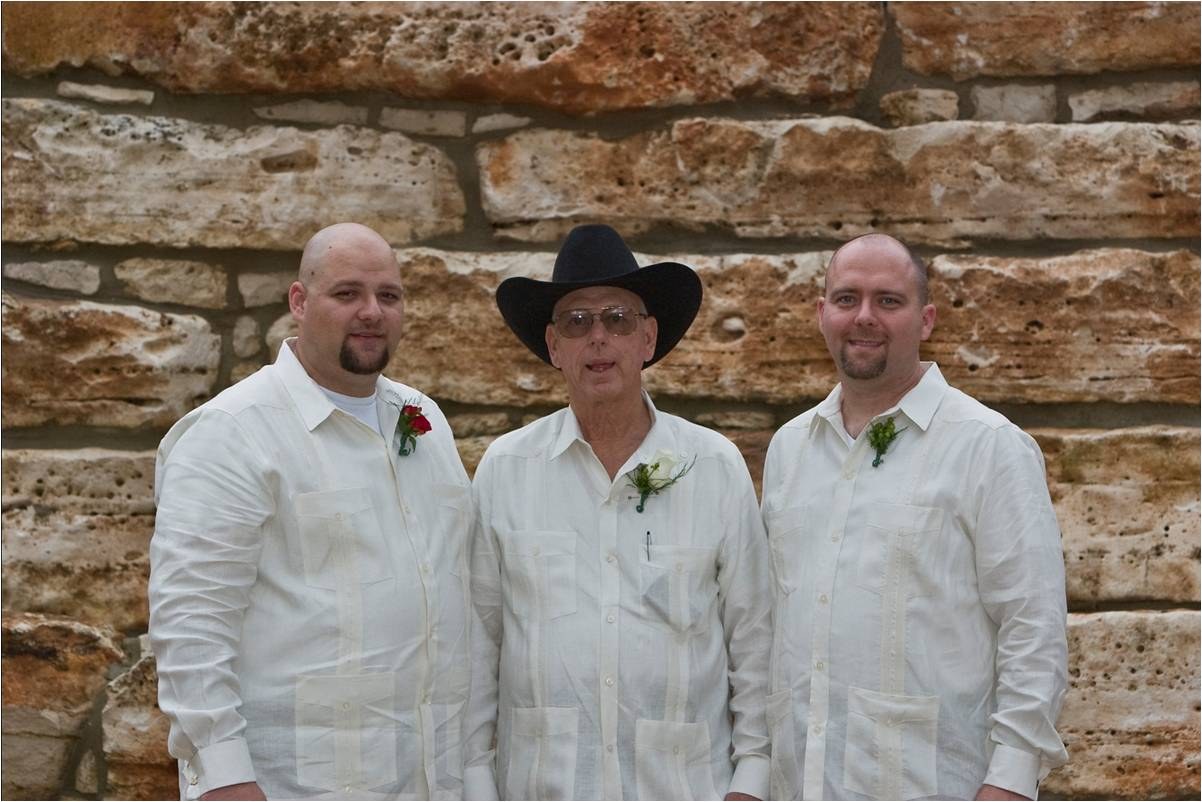 Three men in white shirts with boutonnieres before a stone wall, the one in the middle in a black hat.