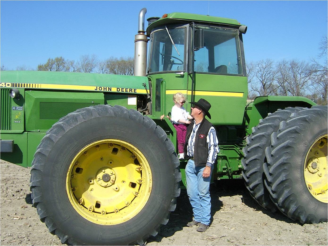 A man in a hat and vest by the front wheel of a big green John Deere tractor, a woman on the step behind.